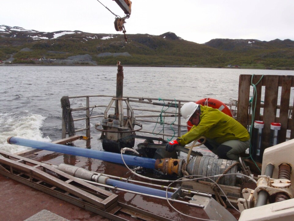 Inspecting a sample taken in the bottom sediment sampler. Two different corers are seen lying on the deck of the Geological Survey of Norway research vessel Seisma. The old processing plant in Repparfjord can be seen in the background. Photo: TE Finne / G
